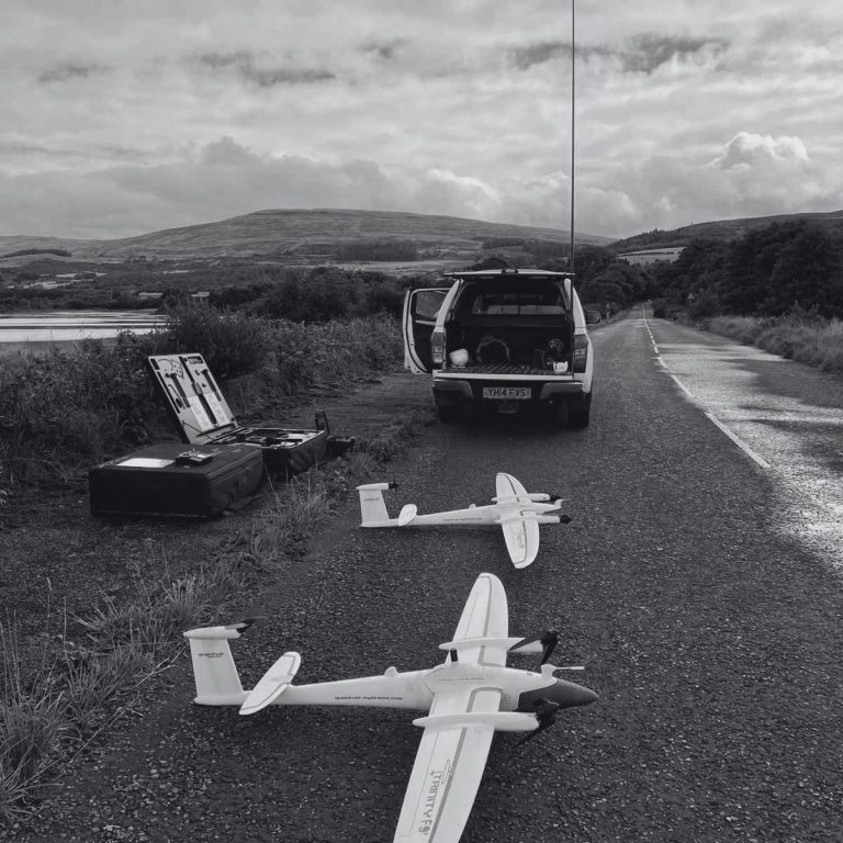 Large-Scale Land Surveys Two fixed-wing drones on a gravel road beside a parked vehicle and a scenic landscape ready to perform a topographic survey in Scotland.