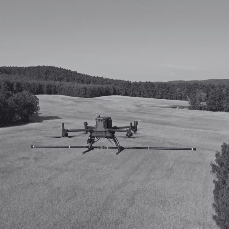 Subsurface Surveys A drone flying over a vast, open field with trees in the background conducting a magnetometer survey for sub-surface anomalies